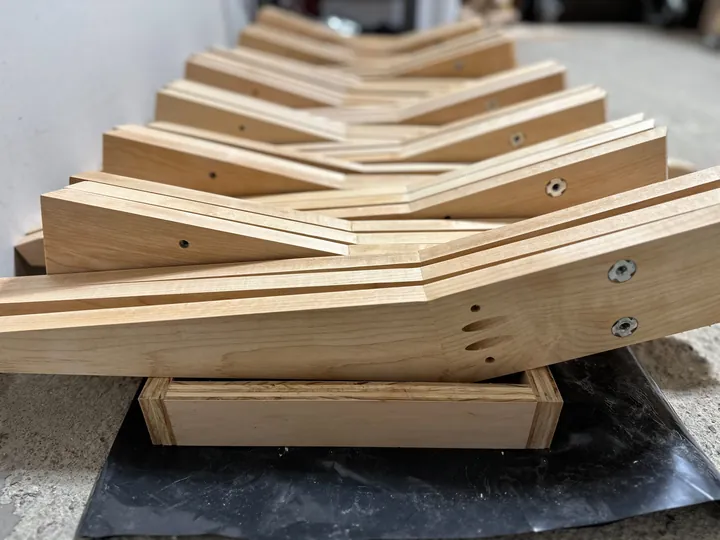 A batch of maple stand parts with screws and T-nuts on a dark workbench.