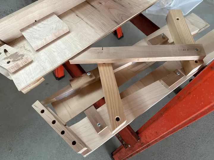 Shaped maple stand parts resting across a wooden jig on the workbench.