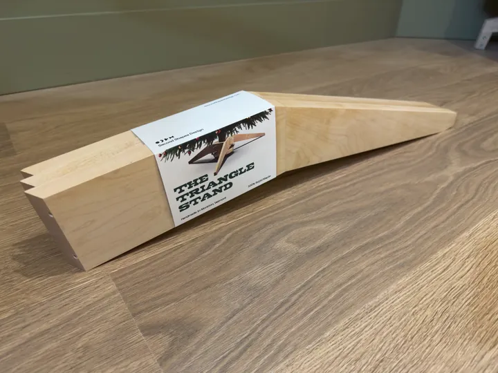 Three maple stand legs packed flat in an illustrated paper wrap on a wooden floor.