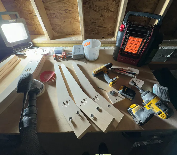 Maple stand parts on the workshop bench among a sander, drill, and hand tools.